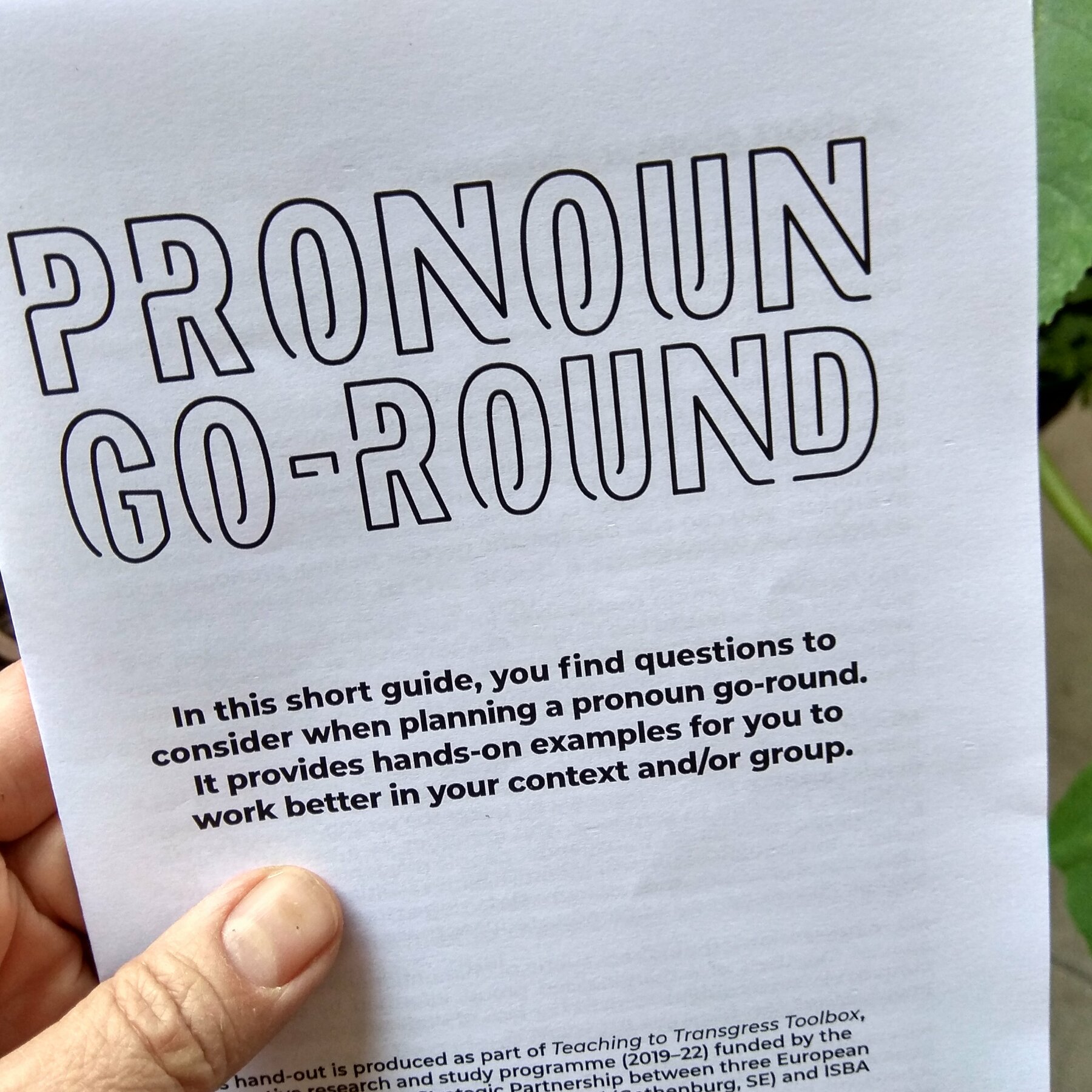A photograph of a hand holding the printed zine *Pronoun Go-Round Guide*.